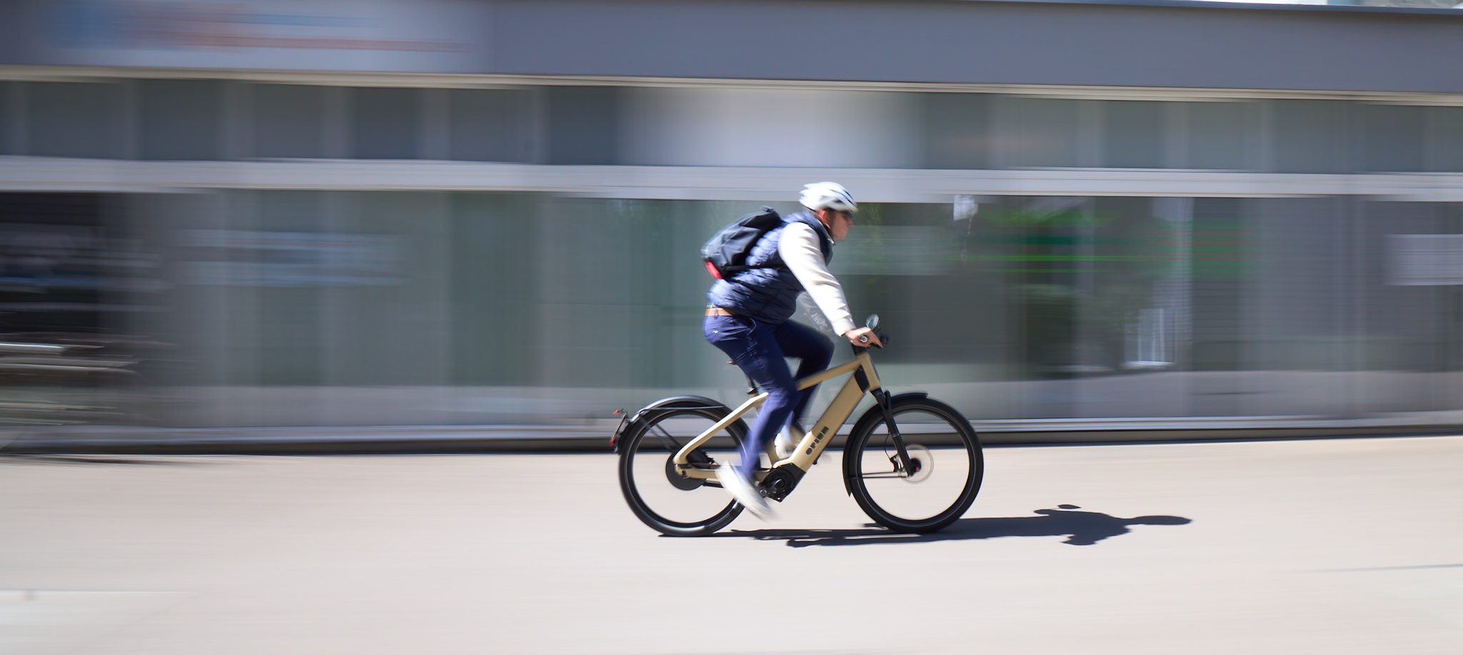 Rider on a Champagne Opium e-bike passing a glass-fronted building, motion-blurred background